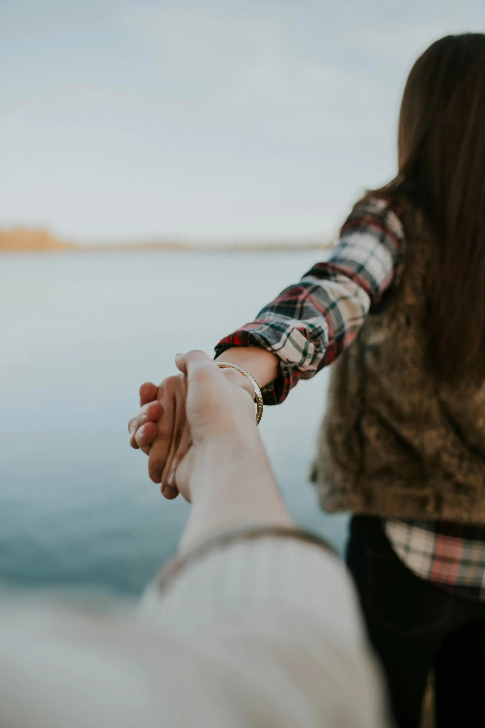 A person with long hair and plaid shirt leads another, holding hands near a calm body of water, evoking a sense of adventure.
