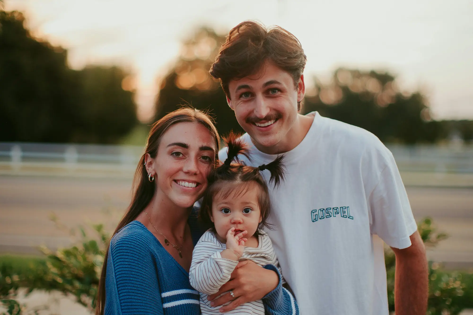 Smiling family outdoors, parents holding a baby, warm sunset background.