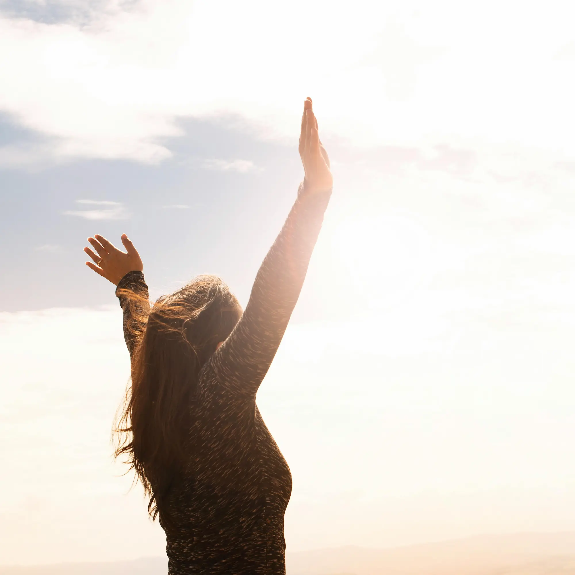 Person with raised arms in sunlight, wearing a long-sleeve shirt, expressing freedom or joy.